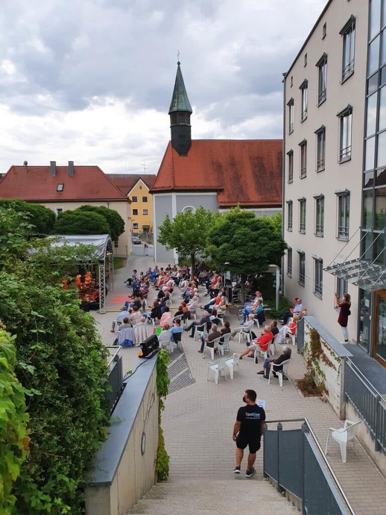 Foto des Spitalgartens mit der Open-Air-Bühne. Zahlreiche Personen sitzen vor einer Bühne. Im Hintergrund ist die Spitalkirche zu sehen.